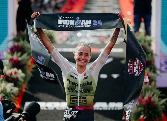 The image shows Laura Philipp crossing the finish line at the 2024 Ironman World Championship. She is smiling and holding up a banner that reads “VinFast Ironman 24 World Championship.” Wearing a race suit with the logos “hepsolar” and “Kickass Sports,” s