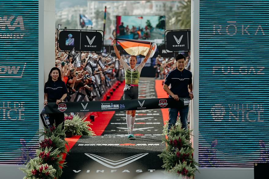 The image shows Laura Philipp crossing the finish line at the 2024 Ironman World Championship, holding a German flag above her head. She is smiling and walking under the banner that reads “VinFast Ironman 24 World Championship.” Spectators are cheering al